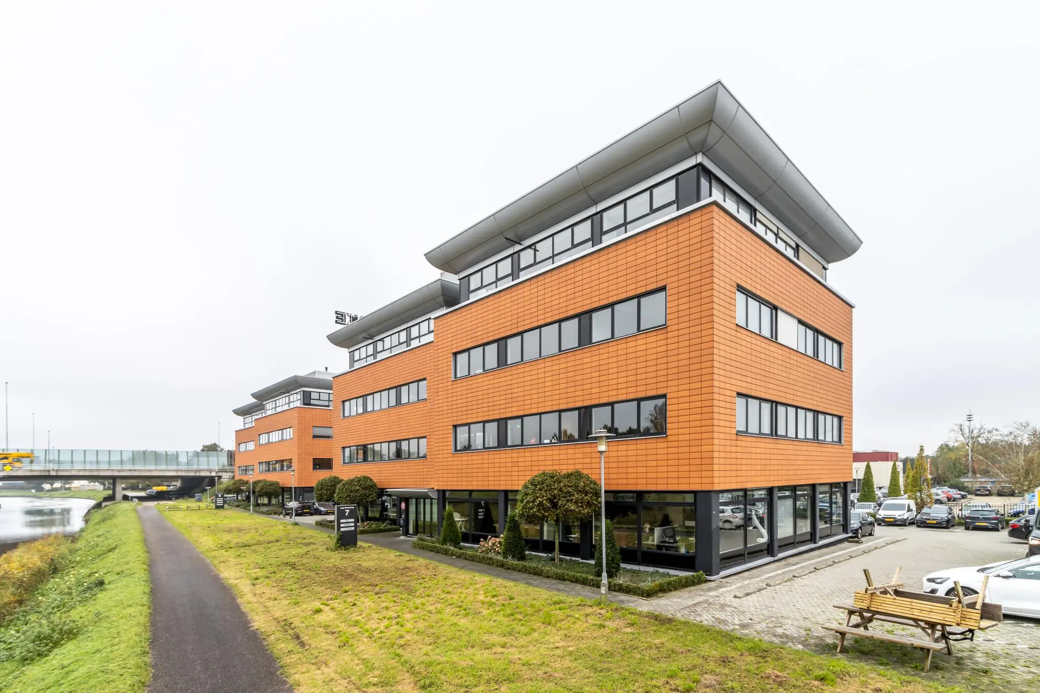 Modern kantoorpand aan de Lage Biezenweg met oranje gevelbekleding en grote ramen, gelegen naast een waterloop en fiets-/wandelpad.